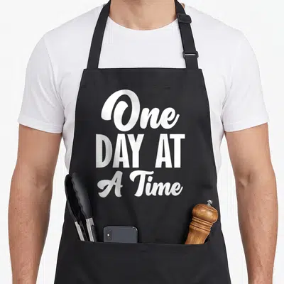 Sobriety addiction recovery apron - White stacked typography reading One DAY AT A Time on grey background — shown on a black cotton canvas apron against a white background