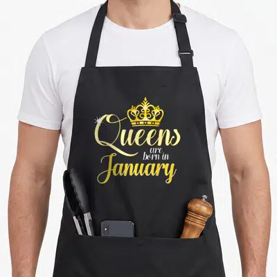 Queen january birthday apron - Yellow crown above stacked script reading Queens are born in January with a white — shown on a black cotton canvas apron against a white background