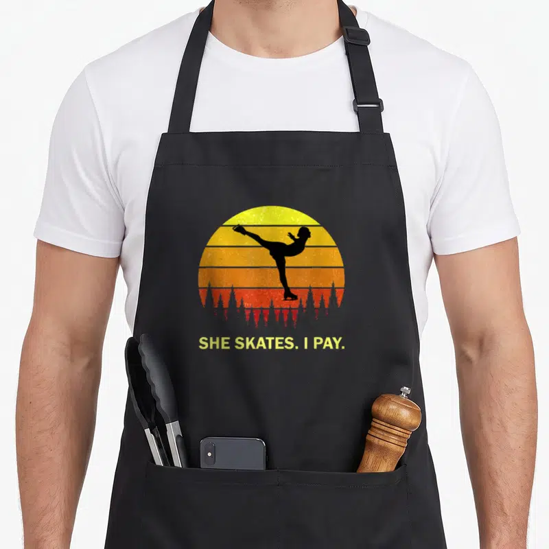 Figure mom daughter apron - Black skater silhouette on yellow orange red striped sun with text SHE SKATES. I PAY — shown on a black cotton canvas apron against a white background