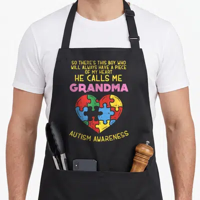 Autism grandma apron - Puzzle piece heart in red blue green and yellow below yellow and pink typography reading So — shown on a black cotton canvas apron against a white background