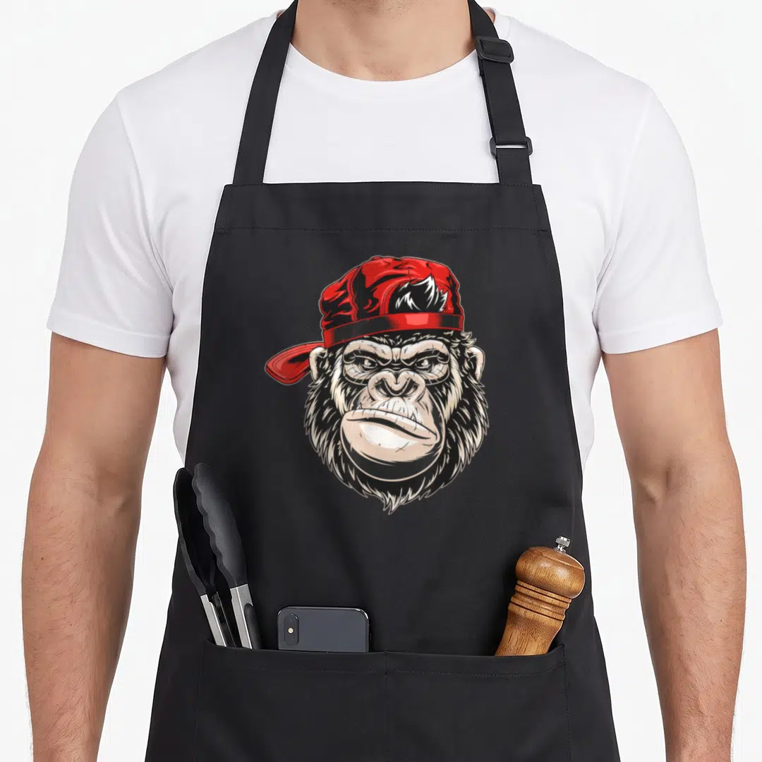 King alpha apron - Cartoon gorilla head wearing a red cap in black white and red ink — shown on a black cotton canvas apron against a white background