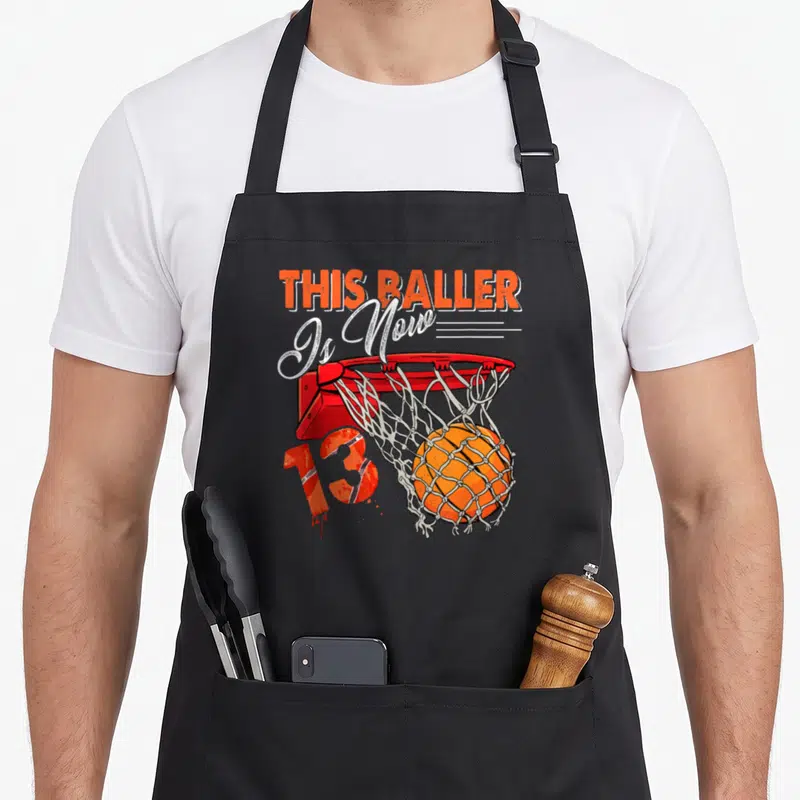 Basketball birthday apron - Cartoon hoop with ball and THIS BALLER Is Now 13 text — shown on a black cotton canvas apron against a white background