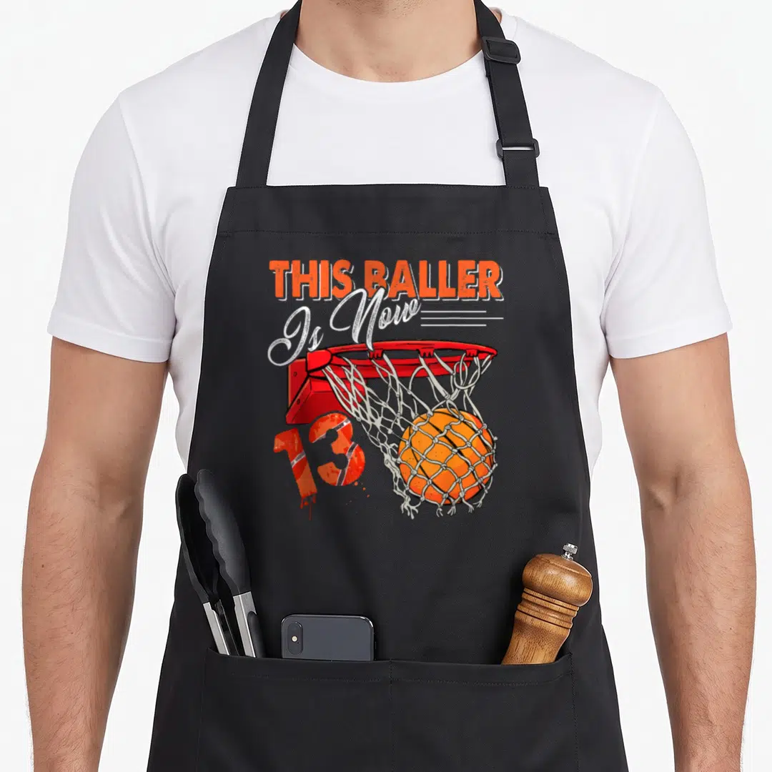 Basketball birthday apron - Cartoon hoop with ball and THIS BALLER Is Now 13 text — shown on a black cotton canvas apron against a white background