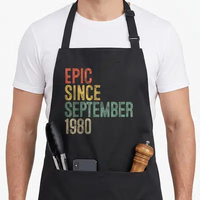 Epic 40th birthday apron - Stacked distressed text reading EPIC SINCE SEPTEMBER 1980 in orange, yellow, green, and beige — shown on a black cotton canvas apron against a white background