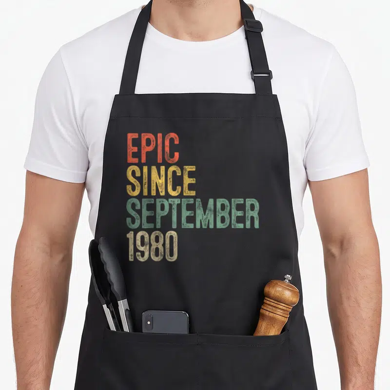 Epic 40th birthday apron - Stacked distressed text reading EPIC SINCE SEPTEMBER 1980 in orange, yellow, green, and beige — shown on a black cotton canvas apron against a white background
