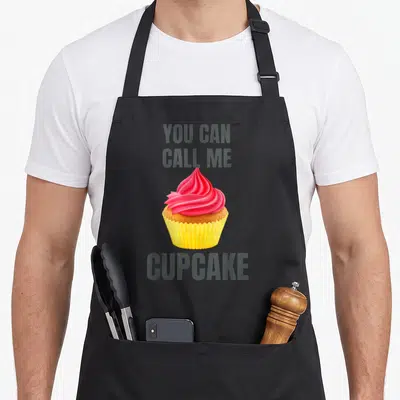 Funny cupcake apron - Cartoon cupcake with pink frosting and grey You Can Call Me Cupcake text — shown on a black cotton canvas apron against a white background