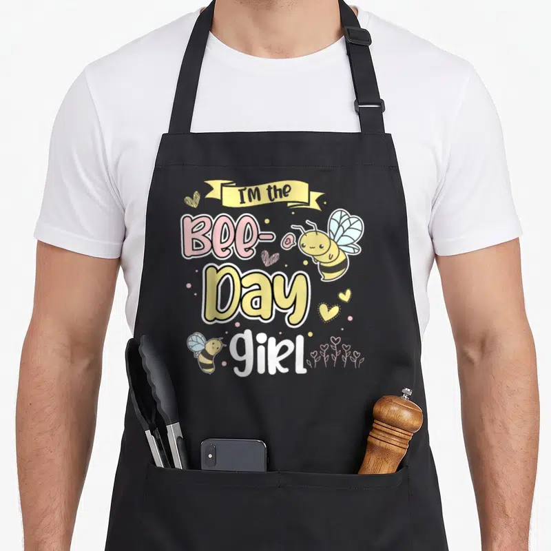 Cute birthday bee aprons - Cartoon bees and hearts surround stacked text reading I'M the Bee-Day girl in yellow pink — shown on a black cotton canvas apron against a white background