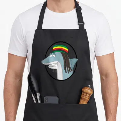 Shark reggae apron - Cartoon blue shark wearing a green yellow red and black striped hat with brown dreadlocks inside — shown on a black cotton canvas apron against a white background