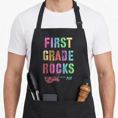 Rockstar teacher apron - Stacked FIRST GRADE ROCKS text above horizontal distressed electric guitar in pink blue green — shown on a black cotton canvas apron against a white background
