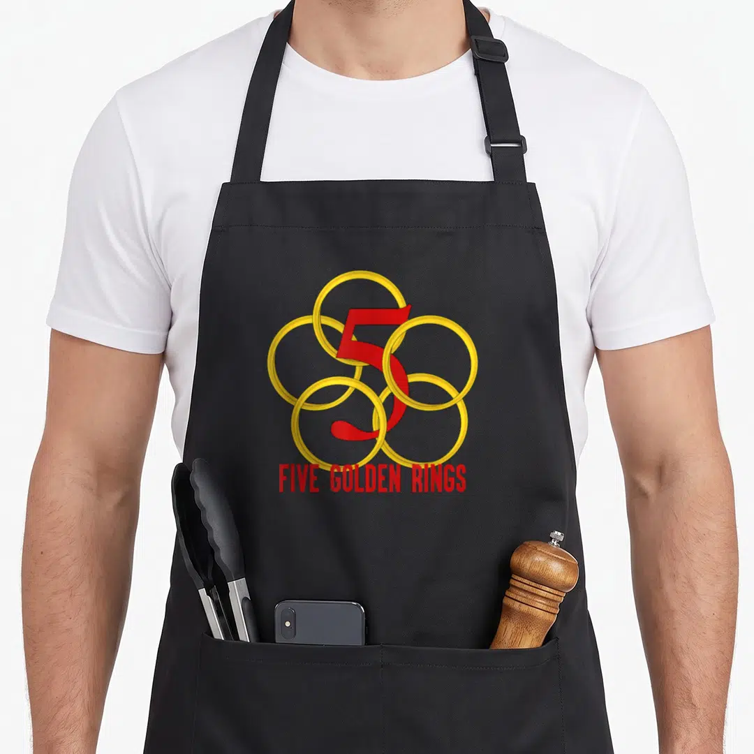 Five christmas apron - Five golden rings interlaced cluster with red number 5 and red text below — shown on a black cotton canvas apron against a white background