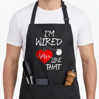 Wired joke apron - Red heart with white EKG line and connected device under stacked distressed text — shown on a black cotton canvas apron against a white background