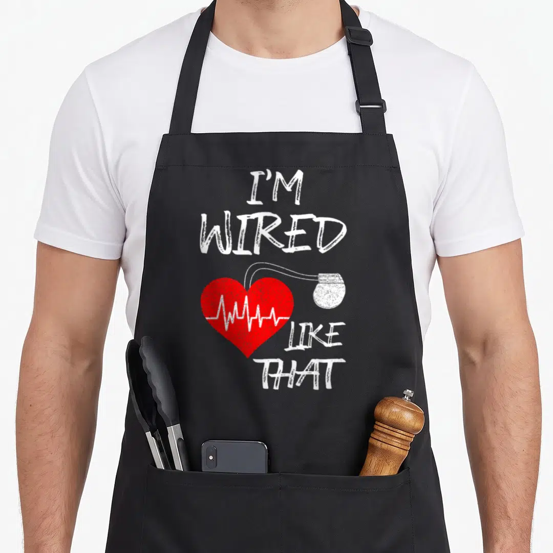 Wired joke apron - Red heart with white EKG line and connected device under stacked distressed text — shown on a black cotton canvas apron against a white background