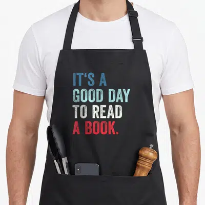 Cute read reading book apron - Stacked text IT'S A GOOD DAY TO READ A BOOK in blue light blue white red — shown on a black cotton canvas apron against a white background