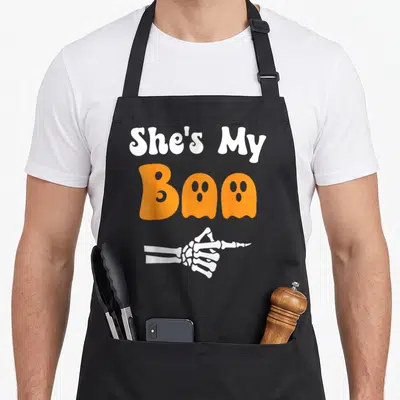 Boo couple apron - White text She's My Boo with orange ghosts and pointing skeleton hand — shown on a black cotton canvas apron against a white background
