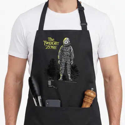 Laser twilight apron - Astronaut with one eye helmet, child looking up, yellow text, grey background, black trees — shown on a black cotton canvas apron against a white background