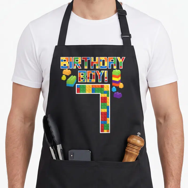 7th building apron - BIRTHDAY BOY! text and number 7 formed from red blue yellow green purple toy bricks with scattered — shown on a black cotton canvas apron against a white background