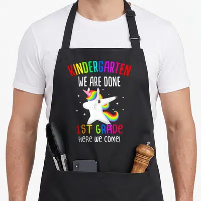 Class unicorn aprons - Dabbing unicorn with rainbow mane and text reading KINDERGARTEN WE ARE DONE 1ST GRADE HERE WE — shown on a black cotton canvas apron against a white background