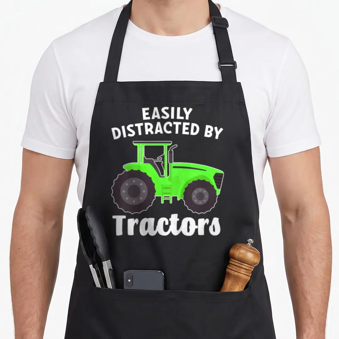 Easily apron or apron - Green cartoon tractor with white text EASILY DISTRACTED BY Tractors on black background — shown on a black cotton canvas apron against a white background