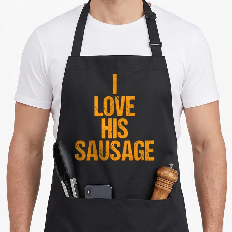Taco sausage apron - Stacked orange text reading I LOVE HIS SAUSAGE in distressed block letters — shown on a black cotton canvas apron against a white background