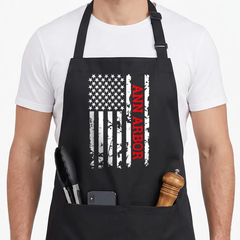 Funny ann arbor apron - Distressed USA flag with vertical red ANN ARBOR text on white background — shown on a black cotton canvas apron against a white background