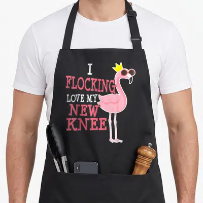 Replacement joke apron - I Flocking Love My New Knee text with crowned flamingo in pink white yellow — shown on a black cotton canvas apron against a white background