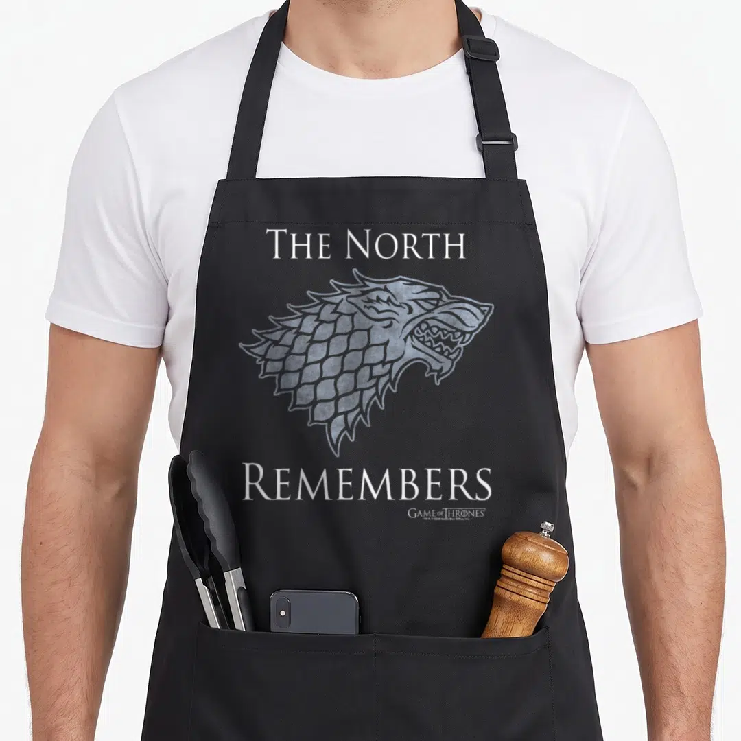 Remember north apron - Grey direwolf head between white THE NORTH REMEMBERS text — shown on a black cotton canvas apron against a white background